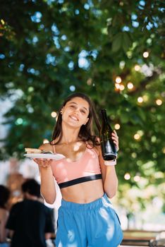A young woman carries a plate of snacks in one hand and a drink in the other