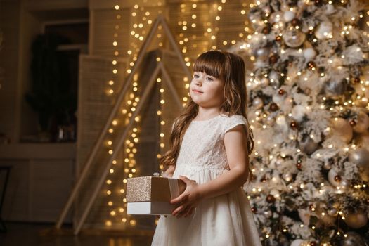 A girl in a festive light dress with a gift in her hands next to a Christmas tree, lights of garlands in the background. The concept of New Year holidays.