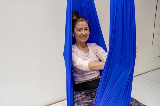 A young woman poses while doing anti-gravity aerial yoga in a blue hammock on a white background