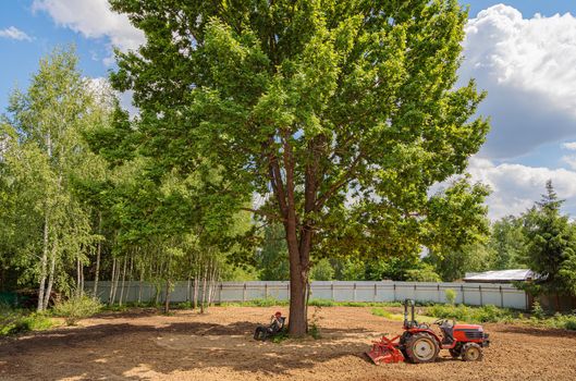 Tractor driver rests in the shade of a tree after plowing the land. Agricultural work on the site.