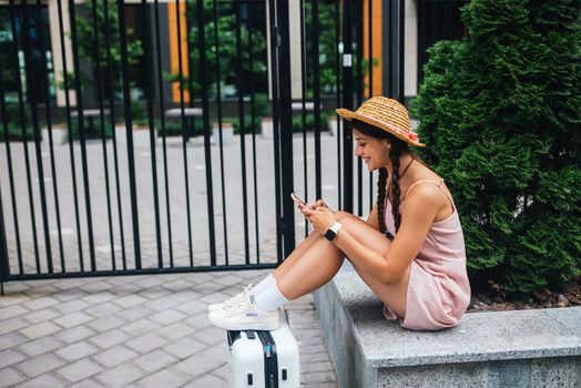 Young slender woman is sitting on a bench with mobile phone, gray suitcase standing by legs, side photo