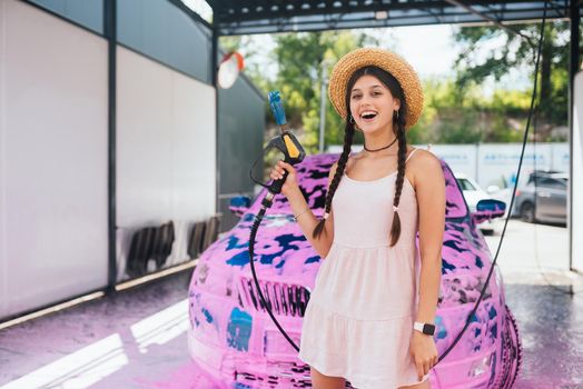 Young woman high pressure hose stands in front of a car covered in pink foam