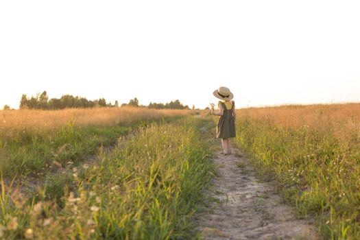 A little blonde girl in a straw hat walks in a field with a bouquet of daisies. The concept of walking in nature, freedom and an eco-friendly lifestyle