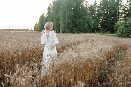 A blonde woman in a long white dress walks in a wheat field. The concept of a wedding and walking in nature.