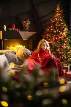 A blonde little girl in a festive outfit next to a Christmas tree decorated with garlands, glass balls and Christmas toys. The concept of winter holidays is Christmas and New Year holidays. Magical festive atmosphere.