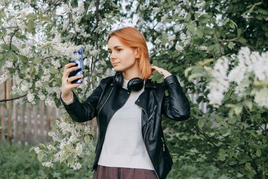 A red-haired teenage girl with headphones around her neck takes a selfie against the background of a flowering cherry tree. The time of spring flowering of cherry orchards.