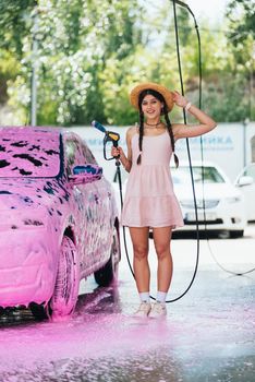 Young woman with high pressure hose stands by car covered in pink foam