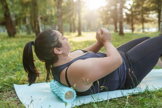 A charming brunette woman plus-size body positive practices sports in nature. Woman is engaged with a massage roller for the body in the park on a sports mat.