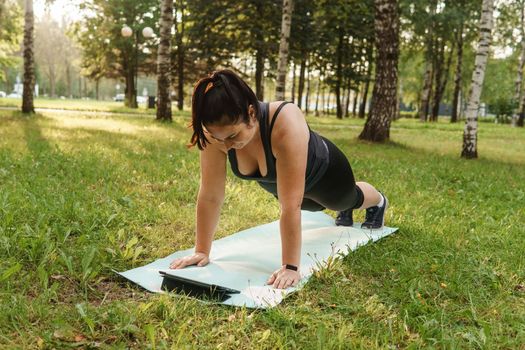 A charming brunette woman plus-size body positive practices sports in nature. Woman does yoga in the park on a sports mat. On a yoga mat in the dog pose.