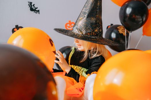 Children's Halloween - a girl in a witch hat and a carnival costume with airy orange and black balloons at home. Ready to celebrate Halloween.