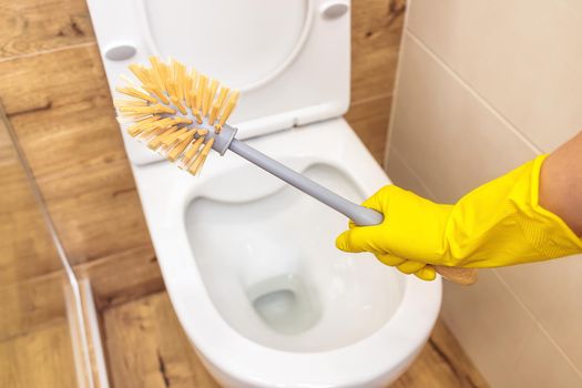 close-up woman's hand in protective yellow glove holds toilet brush to clean the white toilet bowl. The concept of cleaning product, hygiene and disinfection of bathroom at home and in public places