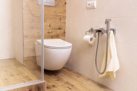 a white toilet and a hygienic shower for a bidet in the bathroom with ceramic tiles in natural colors like wood and white, in a Scandinavian style. Small bathroom interior. Close-up of plumbing.