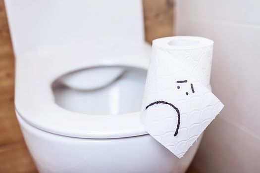 A close-up of a toilet paper roll with a funny emoji stands on a toilet bowl with an open lid in the bathroom. The concept of good health, clean bathroom, happy life