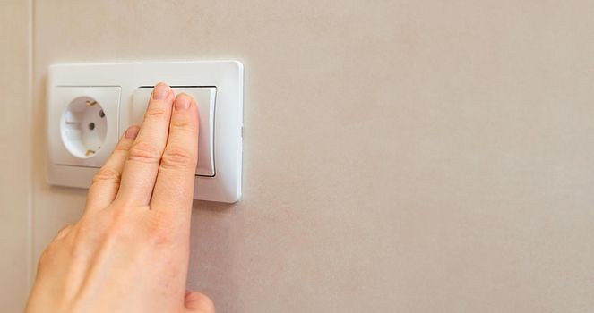 Close-up woman's hand turns on light in the apartment. A white switch along with an electrical outlet on wall background. Copy paste white space for your design. Energy saving and electricity concept