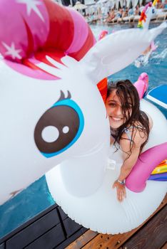 Happy young woman sitting on inflatable unicorn toy mattress by the pool. Girl relaxing sunbathing enjoying travel holidays at resort pool