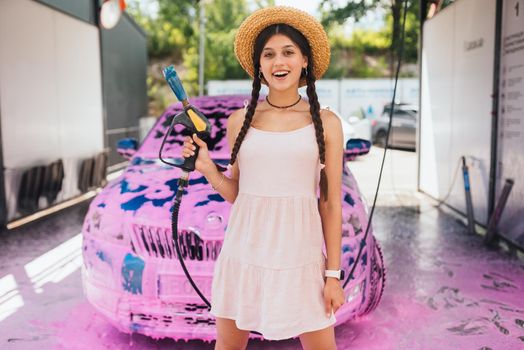 Young woman high pressure hose stands in front of a car covered in pink foam