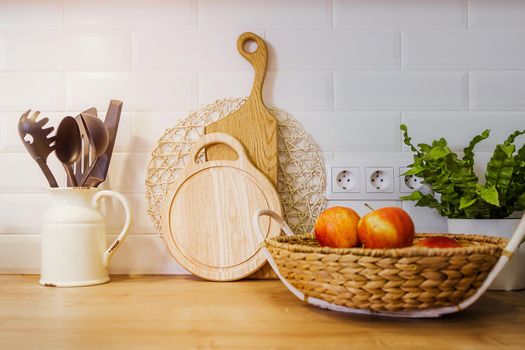 Modern composition mockup in kitchen interior with vegetable cutting boards, apples in a basket, kitchen utensils, home plant in pot and copy space on wooden table.