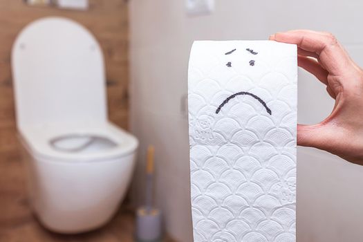 close-up in woman's hand roll of toilet paper on which sad letter is drawn. The concept of health problems, stomach, stomach or hemorrhoids. The background is beautiful bathroom with white toilet bowl