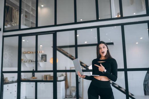 Young woman in a sports suit standing holding a laptop in her hands at home