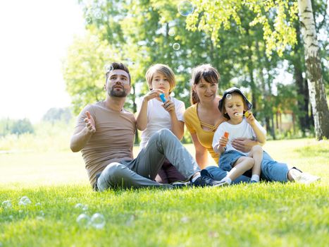 Spending time together. Happy family sitting on grass in summer park and blowing soap bubbles with children