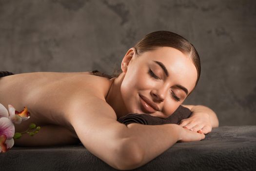 Focus on foreground of relaxing woman resting on massage table with eyes closed in spa salon. Young smiling caucasian girl lay naked. Rest with spa treatment.