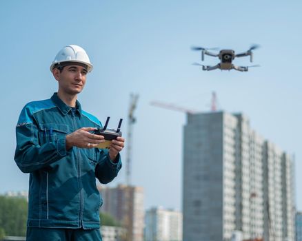 A man in a helmet and overalls controls a drone at a construction site. The builder carries out technical oversight