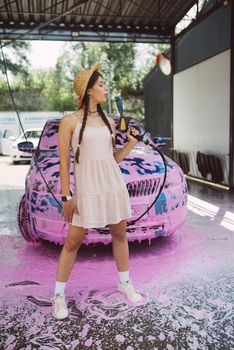 Young woman high pressure hose stands in front of a car covered in pink foam