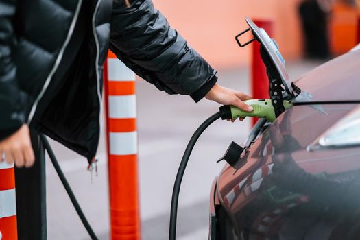 Young beautiful woman traveling by electric car having stop at charging station standing plugging cable browsing internet on smartphone smiling joyful while charing