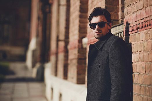 Portrait of fashionable well dressed man with beard posing outdoors looking away, confident and focused mature man in coat standing on a brick wall background, elegant fashion model.