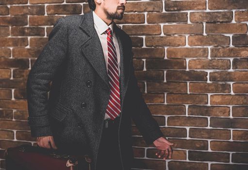 Portrait of fashionable well dressed man with beard posing outdoors looking away, confident and focused mature man in coat standing with suitcase on a brick wall background, elegant fashion model.