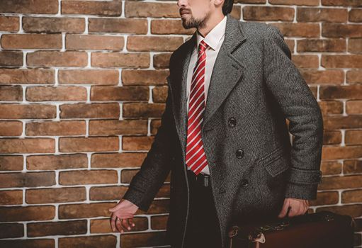 Portrait of fashionable well dressed man with beard posing outdoors looking away, confident and focused mature man in coat standing with suitcase on a brick wall background, elegant fashion model.