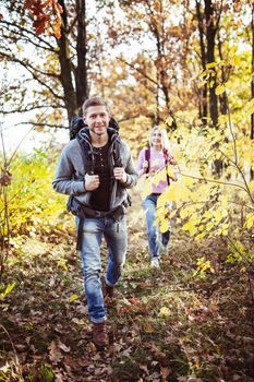 Couple of tourists in love travels in autumn nature. Focus on young Caucasian smiling guy with a big backpack in the foreground, charming blonde follows him on a blurry background.