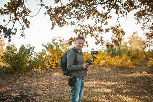 Traveling Operator Prepares Equipment For Shooting, Film Maker Holds Camera On Digital Stabilizer, Caucasian Cameraman Prepare Equipment For Shooting Video Content Standing In Autumn Nature Outdoors