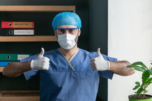 Male Doctor Shows Thumbs Up Sign, Man Wearing Medical Uniform With Disposable Cap And Mask On His Face Raises Hands In Disposable Gloves Showing Thumb Up Sign While Standing In Office