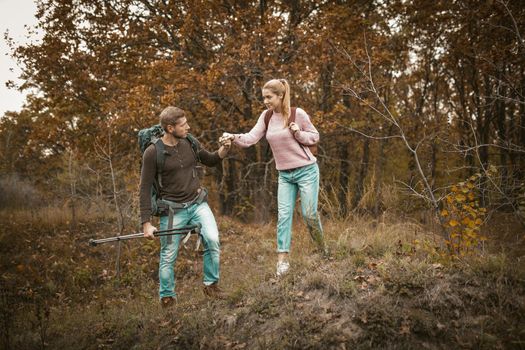 Traveler Man Helps His Woman To Go Down The Hill, Caucasian Man Supports His Woman In Hiking Trip Holding Her Hand And Hiking Sticks In Other Hand Against The Background Of Multicolor Autumn Forest