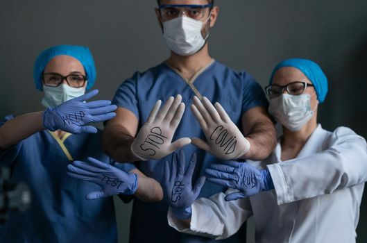 Medics In Protective Uniforms Showing Slogans STOP, HELP, VIRUS, COVID, TEST On Their Hand In Disposable Gloves Pretending To Have Their Hands On Invisible Glass