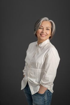 Toothy smiling mature woman stands hands in pockets looking at camera. Gray-haired woman in jeans and white blouse posing on gray background.