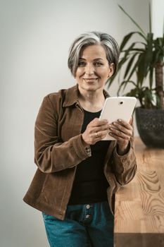 Charming woman uses digital tablet toothy smiling at camera. Mature woman wearing corduroy jacket and jeans holds tablet computer standing near wooden table or sill. Tinted image.