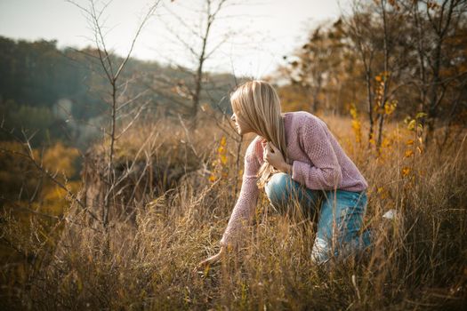 Blonde Looks Into The Distance Crouching On Edge Of Cliff, Caucasian Woman Admires The Beauty Of Sunset Touching Grass With Her Hand, Profile View