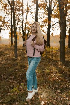 Female traveler posing with a backpack on the background of the autumn forest backlit by the sun's rays. Beautiful blonde travels in nature. Hiking concept.