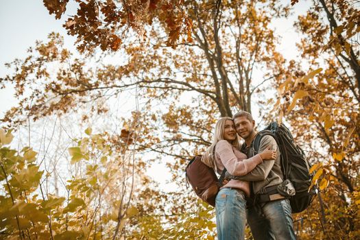 Happy Couple Standing In A Hug Outdoors, Hugging Tourists In Love Stands On Background Of Colorful Autumn Trees And Blue Sky, View From Below