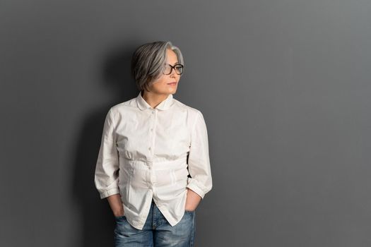 Beautiful mature lady in glasses stands hands in pockets looking at side. Elegant Gray-haired woman in whiite shirt on gray background. Copy space for text at right side.