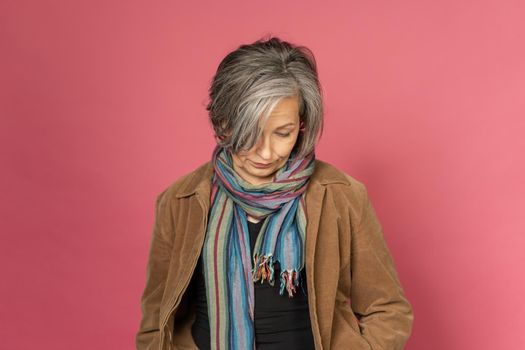 Pensive grey haired woman looks down posing in studio on pink background. Lonely concept.