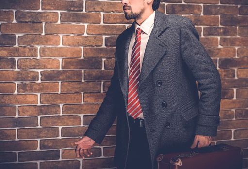 Portrait of fashionable well dressed man with beard posing outdoors looking away, confident and focused mature man in coat standing with suitcase on a brick wall background, elegant fashion model.