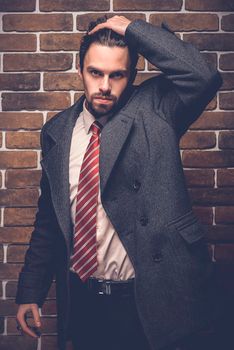 Portrait of fashionable well dressed man with beard posing outdoors looking away, confident and focused mature man in coat standing on a brick wall background, elegant fashion model.