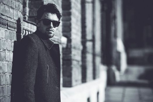 Portrait of fashionable well dressed man with beard posing outdoors looking away, confident and focused mature man in coat standing on a brick wall background, elegant fashion model.