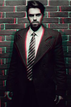 Portrait of fashionable well dressed man with beard posing outdoors looking away, confident and focused mature man in coat standing on a brick wall background, elegant fashion model.