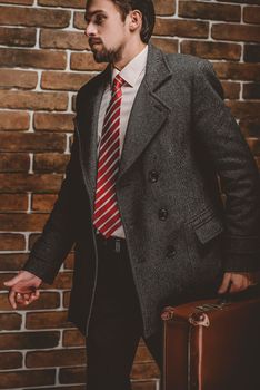 Portrait of fashionable well dressed man with beard posing outdoors looking away, confident and focused mature man in coat standing with suitcase on a brick wall background, elegant fashion model.