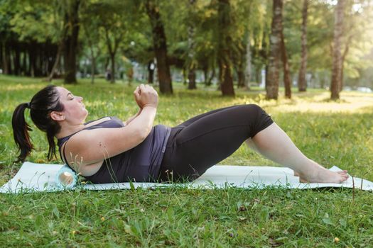 A charming brunette woman plus-size body positive practices sports in nature. Woman is engaged with a massage roller for the body in the park on a sports mat.