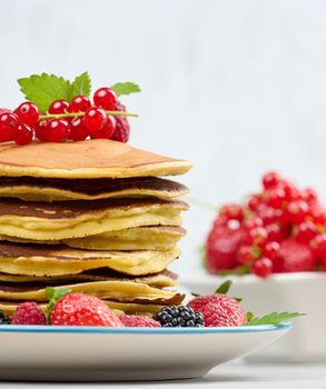 Stack of baked pancakes with fruits in a round plate on a white table